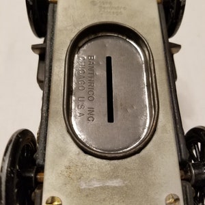 Peut inclure: Tirelire en forme de voiture jouet en m&eacute;tal vintage. Le dessous de la voiture est visible, montrant la fente &agrave; pi&egrave;ces et les marquages du fabricant. Les roues sont noires et le corps est argent&eacute; et gris.