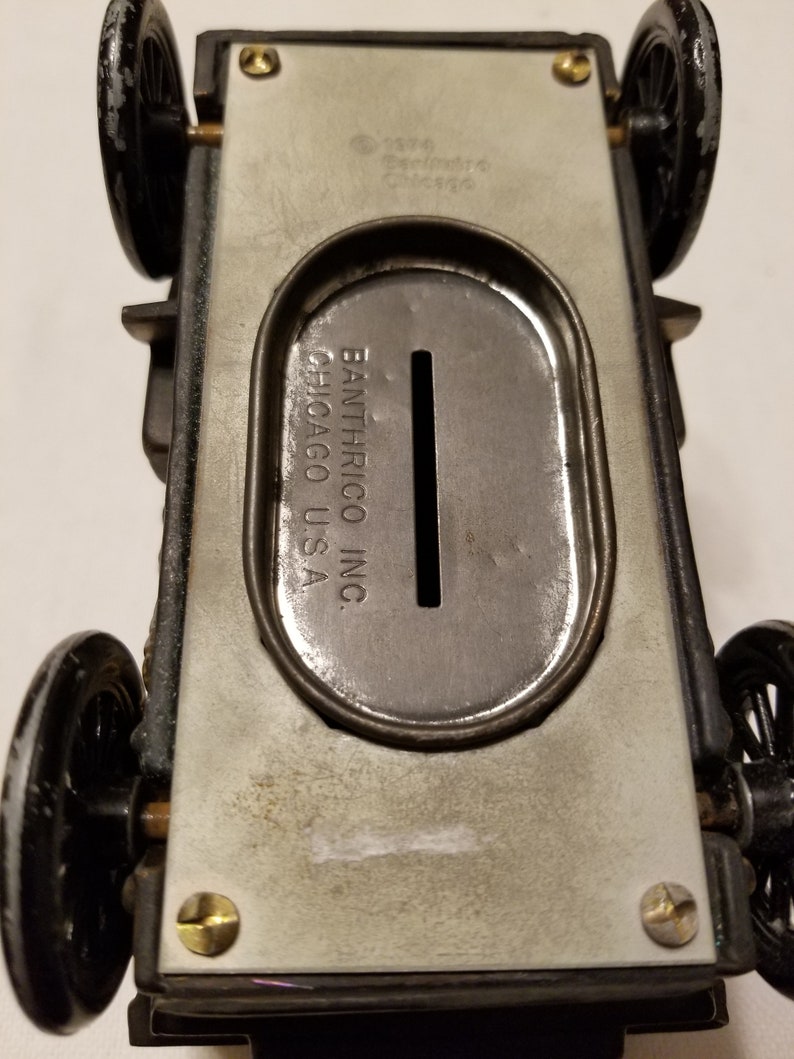 Peut inclure: Tirelire en forme de voiture jouet en m&eacute;tal vintage. Le dessous de la voiture est visible, montrant la fente &agrave; pi&egrave;ces et les marquages du fabricant. Les roues sont noires et le corps est argent&eacute; et gris.