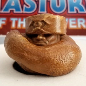 May include: A brown, sculpted figure of a man with a large, round head and a single eye on his forehead. The figure is wearing a brown headband with a raised eye design. The figure is in a seated position with its arms crossed.