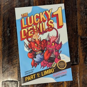 May include: A comic book titled "The Lucky Devils 1: Limbo" features two cartoon devils. One red devil holds a knife, and the other pink devil holds a bottle. The cover has a blue sky background with white clouds.