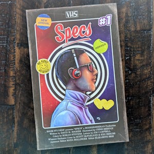 May include: A VHS tape with the title "Specs" and the number "#1" in the upper right corner. The cover art features a person wearing headphones and glasses, set against a red and blue background. Text includes "New Arrival", "Be Kind Rewind", and "Horror".