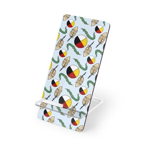 May include: A light blue phone stand with a repeating pattern of feathers, circles, and green vines. The circles are divided into black, white, red, and yellow sections. The stand has a clear plastic base.