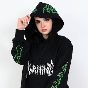 Puede incluir: Sudadera con capucha negra con dise&ntilde;os de llamas verde ne&oacute;n en las mangas y la capucha. La palabra "WARNING" est&aacute; impresa en blanco en la parte delantera. La sudadera tiene una capucha con cord&oacute;n y un bolsillo de canguro.