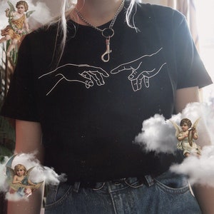 May include: A black t-shirt with a white outline of two hands reaching towards each other. The shirt is worn by a person with blonde hair and a silver necklace with a key charm.