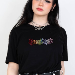 May include: Black t-shirt with the word "FEELINGS" in a rainbow-colored, graffiti-style design. The shirt is paired with black and white checkered pants, a black belt, and a silver necklace.