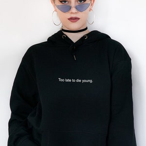 Op de afbeelding: Zwart hoodie met de tekst "Too late to die young." in wit gedrukt op de voorkant.