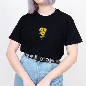May include: A black t-shirt with a small embroidered design of three yellow sunflowers on the front. The model is wearing blue jeans and a black belt with silver grommets.