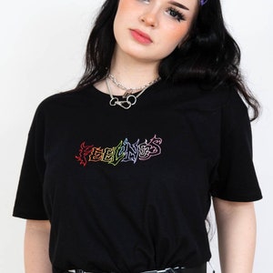 May include: Black t-shirt with the word "FEELINGS" embroidered in rainbow colors. The model is wearing a silver necklace and a black and white checkered belt. The model has black hair and is wearing purple hair clips.