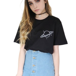 May include: Black t-shirt with a white planet and star design. The shirt is paired with a light blue denim button-up skirt. The model is wearing a black choker necklace.