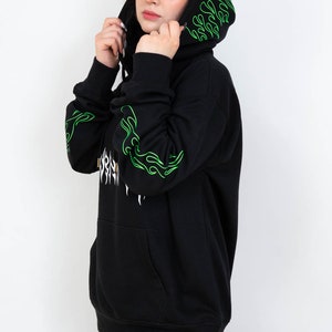 Puede incluir: Sudadera con capucha negra con dise&ntilde;os de llamas verdes en las mangas y la capucha. La sudadera con capucha tiene un bolsillo delantero y un gr&aacute;fico blanco en la parte delantera. La persona lleva un pa&ntilde;uelo negro y blanco en la cabeza.