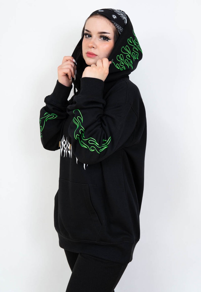 Puede incluir: Sudadera con capucha negra con dise&ntilde;os de llamas verdes en las mangas y la capucha. La sudadera con capucha tiene un bolsillo delantero y un gr&aacute;fico blanco en la parte delantera. La persona lleva un pa&ntilde;uelo negro y blanco en la cabeza.