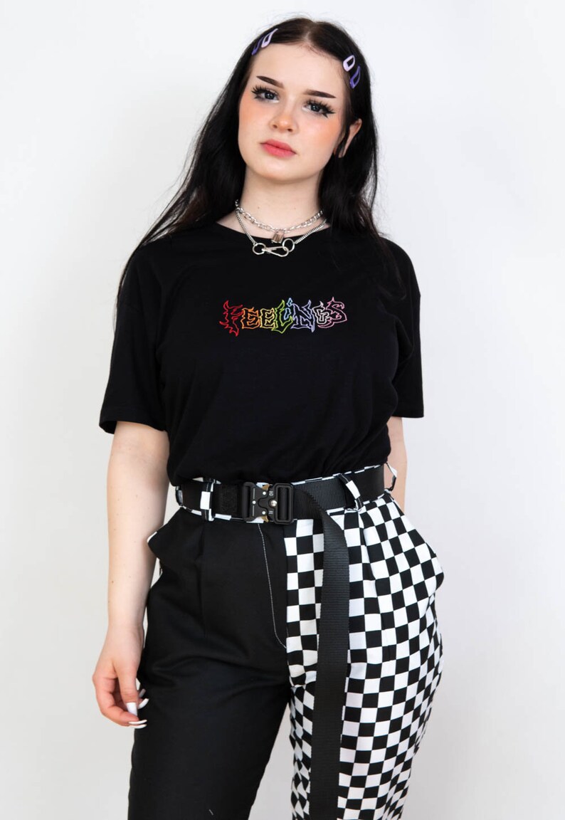 May include: Black t-shirt with the word "FEELINGS" in a rainbow-colored, graffiti-style design. The shirt is paired with black and white checkered pants, a black belt, and a silver necklace.