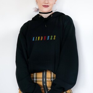 May include: Black hooded sweatshirt with the word "KINDNESS" embroidered in rainbow colors. The model is wearing a yellow and black plaid skirt.