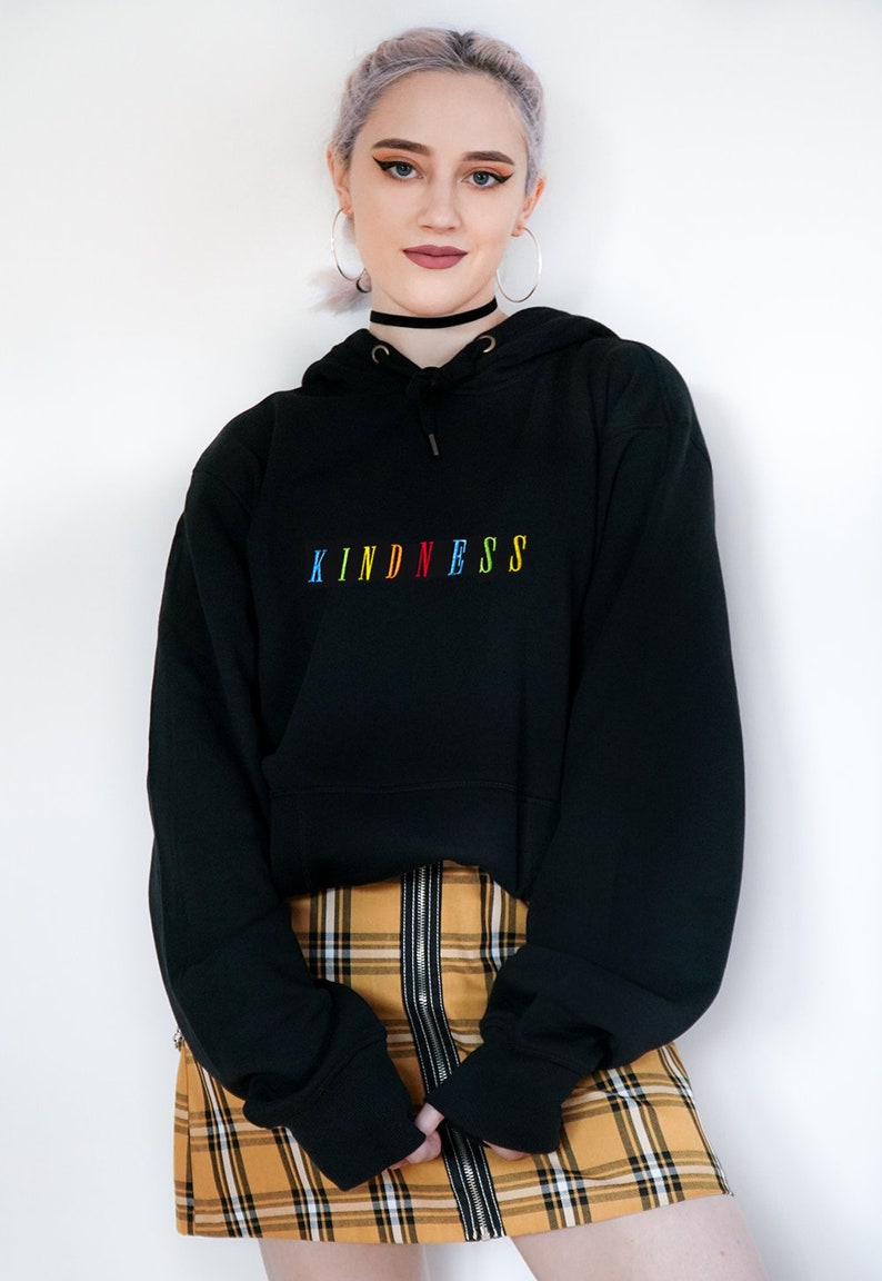 May include: Black hooded sweatshirt with the word "KINDNESS" embroidered in rainbow colors. The model is wearing a yellow and black plaid skirt.