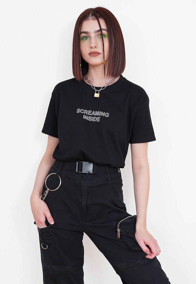 Pode incluir: Uma camiseta preta com o texto "SCREAMING INSIDE" em letras brancas com contorno. A modelo est&aacute; usando cal&ccedil;as cargo pretas com uma corrente prateada e um colar com um cadeado prateado.