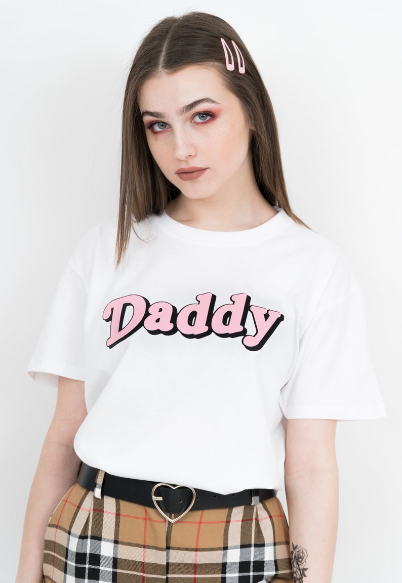 May include: White t-shirt with a pink "Daddy" graphic. The shirt is worn by a person with brown hair and a black belt with a heart-shaped buckle.