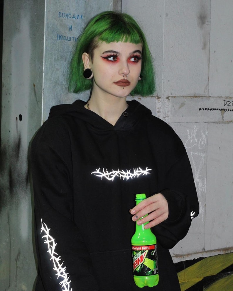May include: A person with short green hair wearing a black hoodie with reflective barbed wire accents. The hoodie has a pocket on the front. The person is holding a green bottle of Mountain Dew with the text "Original Citrus" in Russian.
