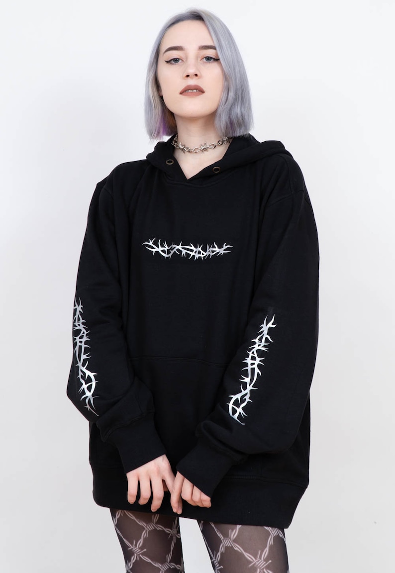 May include: Black oversized hoodie with a silver barbed wire design on the front and sleeves. The hoodie has a drawstring hood and a kangaroo pocket.