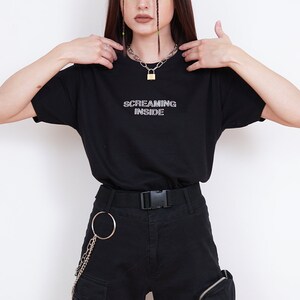 Pode incluir: Uma camiseta preta com o texto "SCREAMING INSIDE" em branco. A modelo est&aacute; usando cal&ccedil;as cargo pretas com uma corrente prateada e um colar com um cadeado prateado.