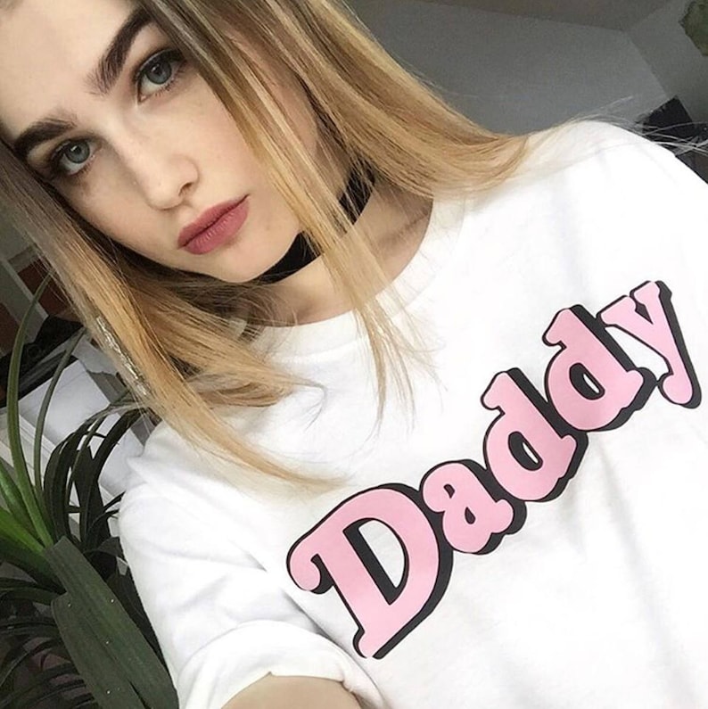 May include: A white t-shirt with a pink "Daddy" graphic printed on the front. The shirt has a black choker necklace around the neck.