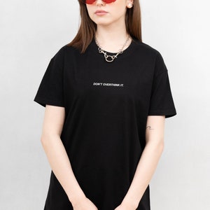 May include: Black t-shirt with the text "DON'T OVERTHINK IT." The shirt is paired with red sunglasses and a silver necklace. The overall style is casual and modern.
