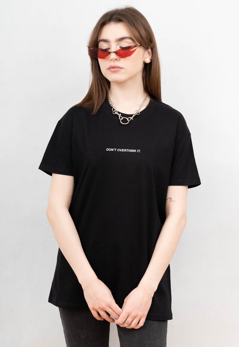 May include: Black t-shirt with the text "DON'T OVERTHINK IT." The shirt is paired with red sunglasses and a silver necklace. The overall style is casual and modern.