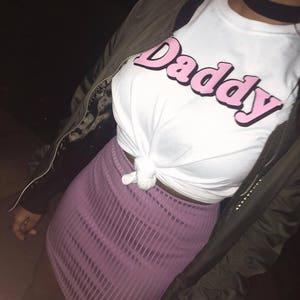 May include: A white t-shirt with a pink "Daddy" graphic. The shirt is tied at the waist and worn with a purple skirt.
