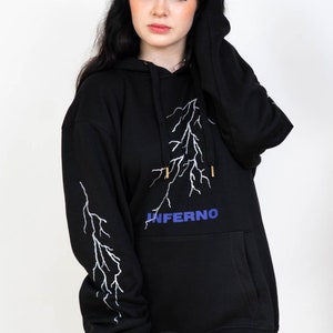 Puede incluir: Sudadera con capucha negra con un dise&ntilde;o de rayo blanco y la palabra "INFERNO" en letras azules.