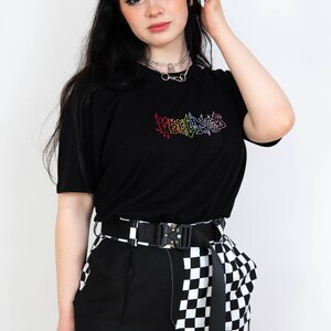 May include: Black t-shirt with rainbow "FEELINGS" text. The model wears a black belt, black and white checkered pants, and a silver necklace. The model has black hair with purple barrettes.