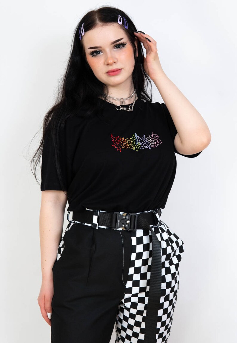 May include: Black t-shirt with rainbow "FEELINGS" text. The model wears a black belt, black and white checkered pants, and a silver necklace. The model has black hair with purple barrettes.