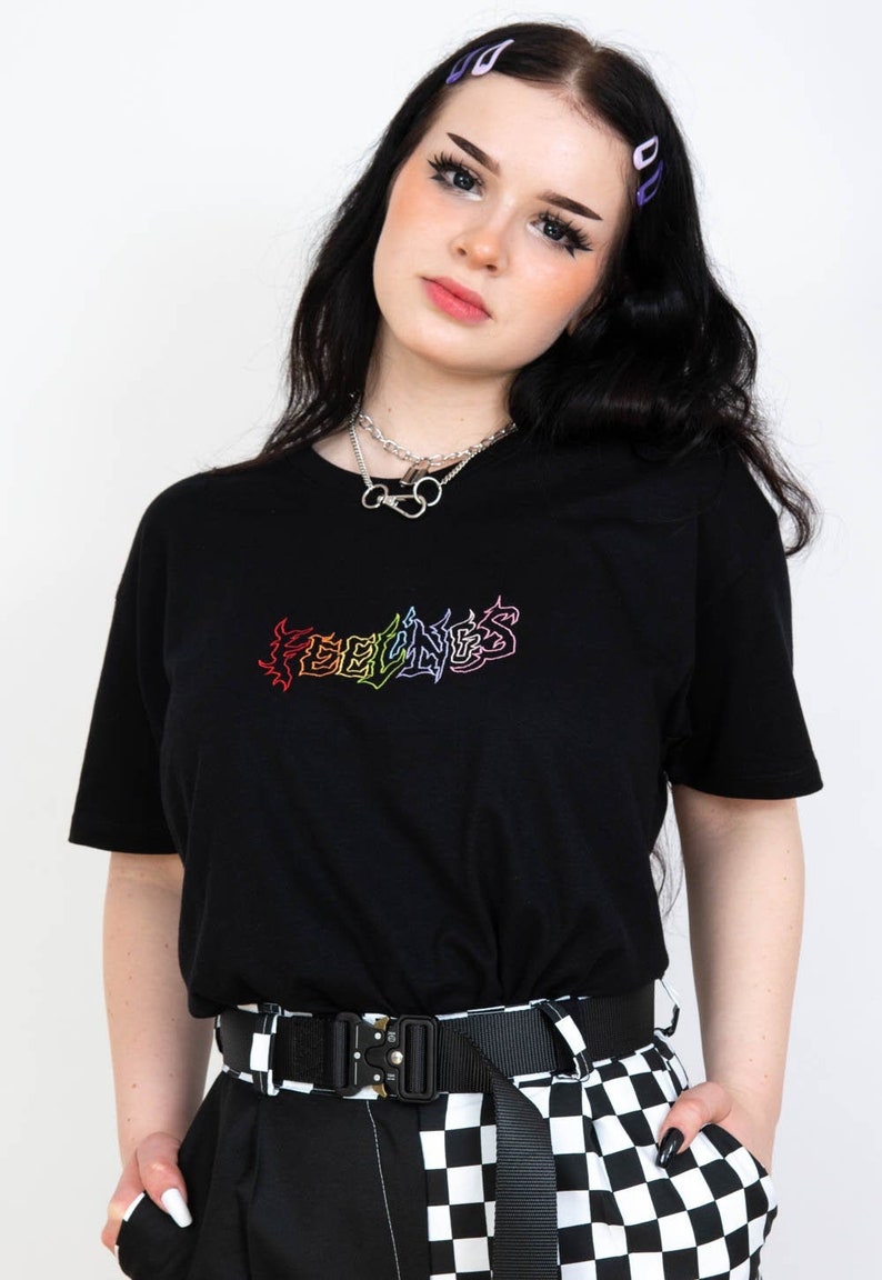 May include: Black t-shirt with the word "FEELINGS" embroidered in rainbow colors. The model is wearing a silver necklace and a black and white checkered belt. The model has black hair and is wearing purple hair clips.