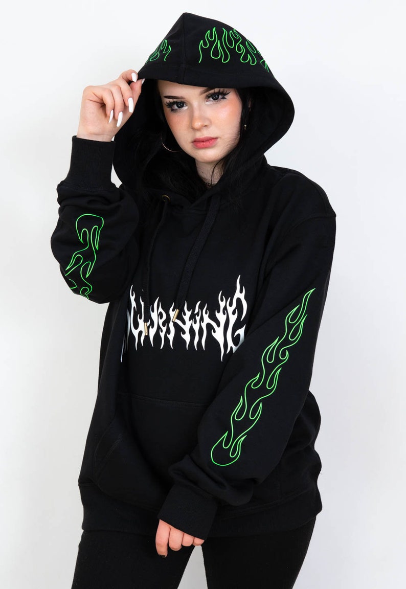 Puede incluir: Sudadera con capucha negra con dise&ntilde;os de llamas verde ne&oacute;n en las mangas y la capucha. La palabra "WARNING" est&aacute; impresa en blanco en la parte delantera. La sudadera tiene una capucha con cord&oacute;n y un bolsillo de canguro.