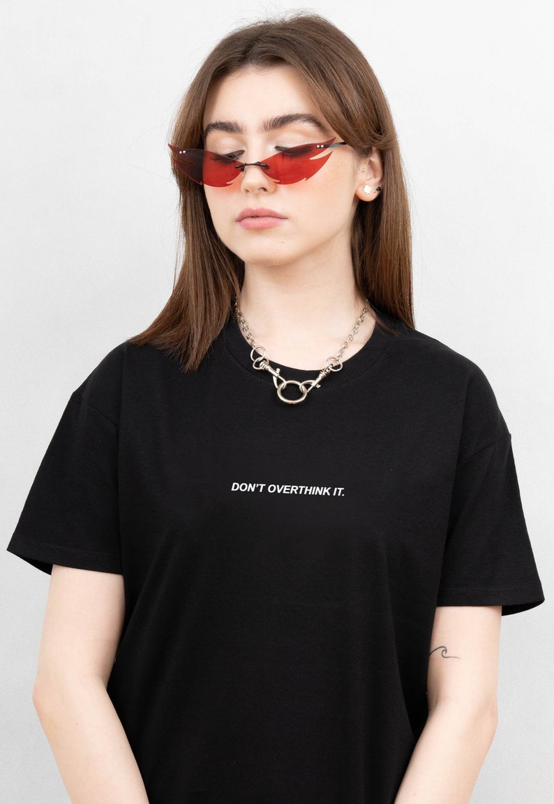 May include: A black t-shirt with the text "DON'T OVERTHINK IT." is paired with a silver chain necklace and red-tinted sunglasses. The sunglasses have a unique, angular shape. The overall style is casual and modern.
