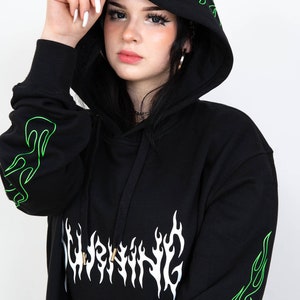 Puede incluir: Sudadera con capucha negra con dise&ntilde;os de llamas verde ne&oacute;n en la capucha y las mangas. La palabra "WARNING" est&aacute; impresa en blanco, con letras en forma de llama en el pecho. La sudadera tiene un cord&oacute;n y un bolsillo tipo canguro.