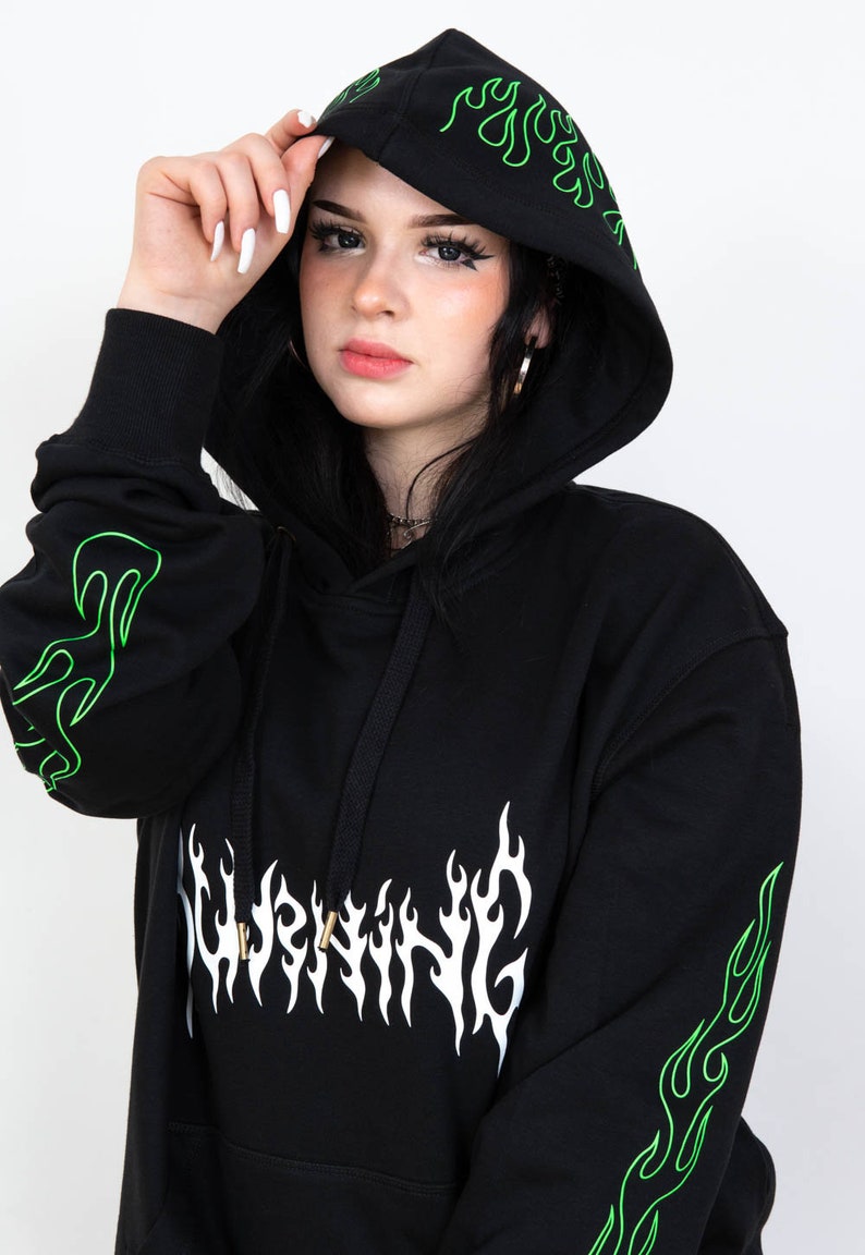 Puede incluir: Sudadera con capucha negra con dise&ntilde;os de llamas verde ne&oacute;n en la capucha y las mangas. La palabra "WARNING" est&aacute; impresa en blanco, con letras en forma de llama en el pecho. La sudadera tiene un cord&oacute;n y un bolsillo tipo canguro.