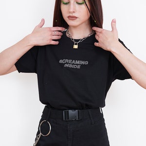 Pode incluir: Uma camiseta preta com o texto "SCREAMING INSIDE" em letras brancas delineadas. A modelo est&aacute; usando um colar de corrente prateada com um pingente de cadeado dourado e cal&ccedil;as cargo pretas com uma corrente e um anel prateados.