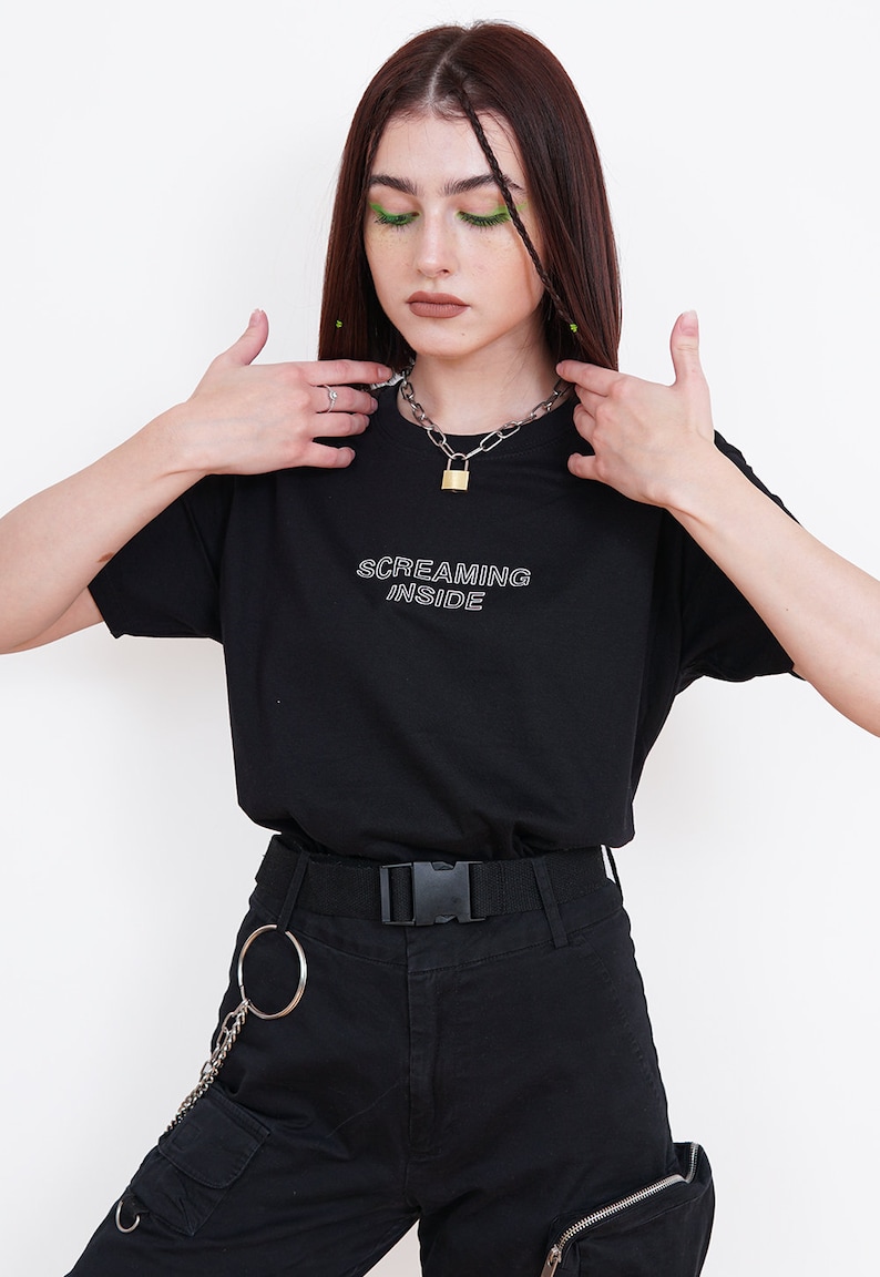 Pode incluir: Uma camiseta preta com o texto "SCREAMING INSIDE" em letras brancas delineadas. A modelo est&aacute; usando um colar de corrente prateada com um pingente de cadeado dourado e cal&ccedil;as cargo pretas com uma corrente e um anel prateados.
