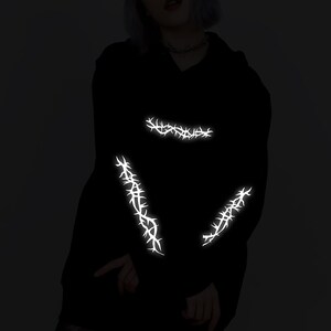 May include: Black hoodie with reflective white thorn designs on the sleeves and chest.