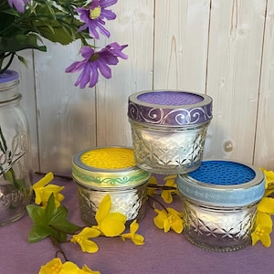 May include: Three glass jars with decorative lids. The lids are purple, yellow, and blue. The jars are filled with white material. The jars are sitting on a purple surface.