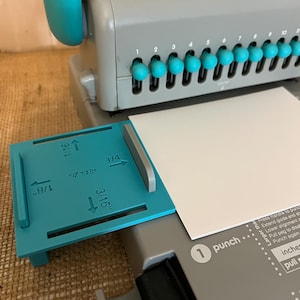 May include: A gray and teal The Cinch book binding machine with a teal alignment guide featuring measurements of 1/16 inch, 1/8 inch, 3/16 inch, and 1/4 inch. The machine has a series of teal punch knobs and a white paper sheet inserted for binding.