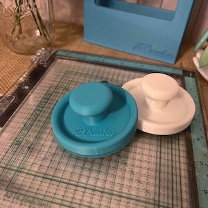 May include: Two stamp press tools, one teal and one white, sit on a grid surface. The teal tool has the text "AF Creates" on its side. A light blue frame is in the background, also with the text "AF Creates".