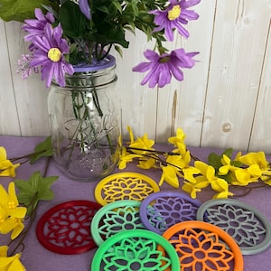 May include: Six colorful 3D printed coasters with a flower design. The coasters are in shades of red, green, yellow, orange, purple, and gray. The coasters are on a purple surface.