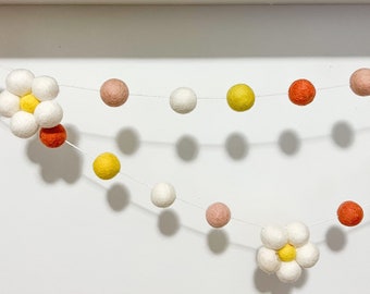 May include: A white felt garland with white, yellow, orange, and pink pom poms and two white felt flowers with yellow centers.