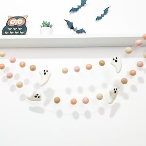 May include: A Halloween garland with white felt ghosts and peach and brown felt balls. The garland is hanging on a white shelf with a black owl figurine and a small potted plant.