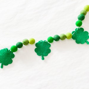 May include: A green felt shamrock garland with green felt balls strung on a white string. The garland is hanging on a white background.