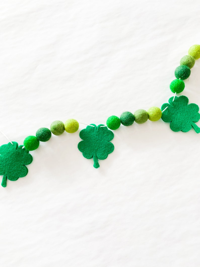 May include: A green felt shamrock garland with green felt balls strung on a white string. The garland is hanging on a white background.