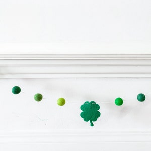 May include: A green felt shamrock garland with green felt balls strung on white twine. The garland is hanging on a white wall.