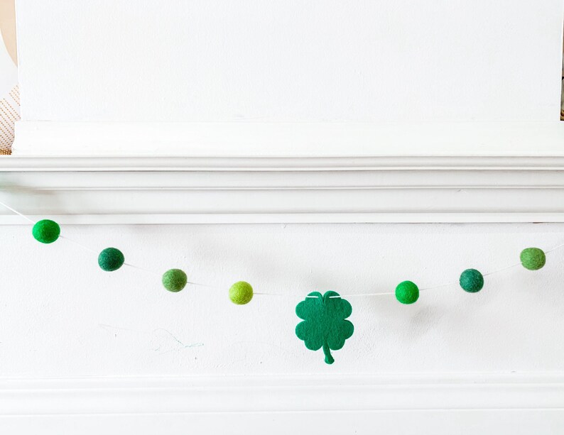 May include: A green felt shamrock garland with green felt balls strung on white twine. The garland is hanging on a white wall.