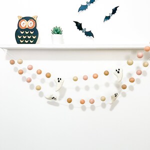 May include: A Halloween garland with white felt ghosts and peach and brown felt balls. The garland is hanging from a white shelf with a wooden owl and a small potted plant.
