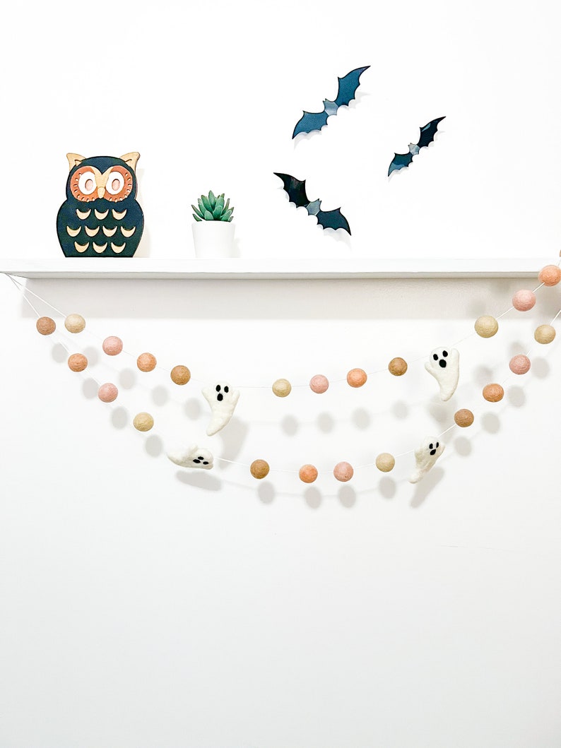 May include: A Halloween garland with white felt ghosts and peach and brown felt balls. The garland is hanging from a white shelf with a wooden owl and a small potted plant.